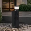 A black outdoor lamp post with two outlets and a motion sensor, placed on a stone patio next to a building. Produktbild-Vorschau