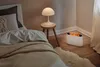 A bedroom scene with a bed, a nightstand with a lamp, and a white electric heater with a flame effect. Produktbild-Vorschau