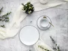 A flat lay of a white plate, a white teacup and saucer with a gold spoon, a small vase with greenery, and a draped white cloth on a textured gray and white surface. Náhľad obrázku produktu