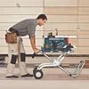 A man is using a portable table saw on a stand with wheels in what appears to be a workshop or construction site. Voir les images du produit