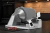 A silver meat slicer is shown on a dark countertop, with sliced meat in front of it. The background shows kitchen cabinets and appliances. Anteprima dell’immagine prodotto