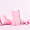 A pink teddy bear keychain with a heart on its chest is displayed on a pink background with other pink decorative items. Podgląd zdjęcia produktu