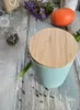 A turquoise ceramic storage jar with a wooden lid sits on a rustic wooden table, surrounded by scattered peppercorns and a sprig of green herbs. Other items like a small glass jar with spices, a pumpkin, and dried flowers are visible in the background. Náhľad obrázku produktu