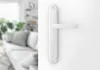 A white door handle is shown on a white background, with a blurred living room in the background. Produktbild-Vorschau