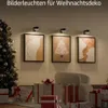 Drei schwarze Bildleuchten, die gerahmte Kunst an einer Wand beleuchten, mit einem Weihnachtsbaum und verpackten Geschenken in der Nähe Náhled obrázku produktu