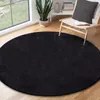 A round black rug is placed on a wooden floor in a room with a large window and a chair. Produktbild-Vorschau