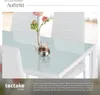 A close-up of a dining table with a glass top, set with a vase of flowers and a candle holder. Chairs are visible in the background. Produktbild-Vorschau