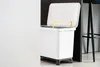 A white trash can with a yellow handle and black legs is shown in a room with a white wall and a black background. Náhľad obrázku produktu