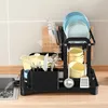 A black dish rack filled with kitchenware is placed on a wooden countertop next to a sink with water flowing into it. Produktbild-Vorschau