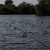 Three fishing rods with orange fishing line are set up on the edge of a body of water, with trees in the background. Anteprima dell’immagine prodotto