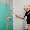 A man is applying a waterproofing compound to a wall with a roller. The wall has green waterproofing material applied to it, and there are plumbing fixtures visible. Produktbild-Vorschau