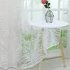 A white sheer curtain with a leaf pattern is draped over a small round table next to a window. A pink rose in a geometric vase sits on the table. Produktbild-Vorschau
