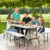 A family is having a picnic in a backyard setting, with a picnic table and benches set up under trees. Produktbild-Vorschau