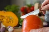 A hand is using a knife to cut a tomato, with a pumpkin, garlic, and other vegetables in the background. Voir les images du produit