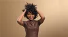 A woman with curly hair is smiling and touching her hair, with a plain brown background. Voir les images du produit