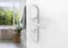 A close-up of a silver door handle and lock mechanism on a white door, with a blurred background of a bathroom. Produktbild-Vorschau
