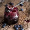 A close-up shot of a jar of blueberry jam with a spoon inside, surrounded by blueberries on a wooden surface. Produktbild-Vorschau
