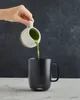 A hand pours green liquid from a small pitcher into a black mug. Náhled obrázku produktu