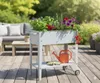 A raised garden bed filled with plants and flowers on a wooden deck, with outdoor furniture in the background. Produktbild-Vorschau