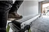A person's leg and boot are stepping onto a long, silver, telescopic plank supported by green legs. The plank is in a construction or workshop environment with concrete floors and white walls. Produktbild-Vorschau