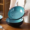 Hands holding a stack of blue and black ceramic bowls on a wooden table. Voir les images du produit