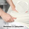 Hands are shown stretching a cream-colored fitted sheet over a mattress, highlighting its elastic edge. Produktbild-Vorschau