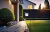 A solar garden lantern illuminates a pathway next to a manicured lawn and hedges, with a house in the background during sunset. Produktbild-Vorschau