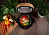 A steaming pot of vegetables on a wooden surface, surrounded by fresh produce and plants. Voir les images du produit