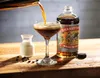 A cocktail is being poured into a coupe glass, with a bottle of Shanky's Whip Original Black Irish Whiskey Liqueur and a small pitcher of cream on a wooden surface. Produktbild-Vorschau