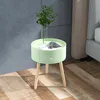 A light green round side table with a drawer and wooden legs, placed next to a bed. A book and a vase with a plant are on top of the table. Voir les images du produit