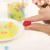 A hand holding a red gel bead with a plate of colorful gel beads in the background. Náhled obrázku produktu