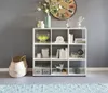 A white cube bookshelf filled with decorative items and books, placed against a gray wall. A woven basket is on the floor next to it. A patterned rug is in the foreground. Produktbild-Vorschau