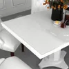A close-up view of a white dining table with a clear protective film on top. Chairs and decor are visible in the background. Produktbild-Vorschau