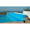 A blue pool cover is being rolled up on a reel next to a swimming pool, with a scenic view of hills and the sea in the background. Produktbild-Vorschau