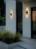 Two modern outdoor wall lights illuminate a building's exterior at dusk, with lush greenery in the foreground. Produktbild-Vorschau