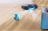 A robot vacuum cleaner is shown navigating a wooden floor, with a teddy bear highlighted by a digital bounding box, indicating object recognition. A sofa is visible on the right. Náhľad obrázku produktu
