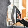 A person is ironing a grey t-shirt with a Black+Decker iron in a home setting. Produktbild-Vorschau