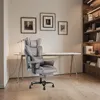 A gray ergonomic office chair is placed in a home office setting with a desk, lamp, and bookshelves. Produktbild-Vorschau