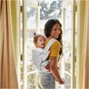 A woman is carrying a baby in a carrier in front of a doorway with curtains. Produktbild-Vorschau