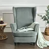 A grey velvet wingback chair with a book and a knitted blanket draped over it, placed next to a woven side table with a lamp. Podgląd zdjęcia produktu