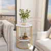 A round side table with two shelves, decorated with a vase of white roses and two bottles, placed between two sofas in a well-lit room with a city view from the window. Produktbild-Vorschau