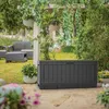 A dark gray Keter storage box is placed on a patio in a garden setting, with plants and outdoor furniture visible in the background. Podgląd zdjęcia produktu