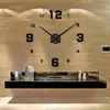 A large, modern wall clock with black numerals and hands is mounted on a light wood-paneled wall above a black floating shelf. The shelf displays a few decorative items. Podgląd zdjęcia produktu