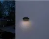An outdoor wall light is illuminated at dusk, casting a warm glow on a textured wall. The background shows a dimly lit outdoor scene with trees and a building. Produktbild-Vorschau