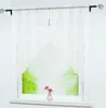 A white lace curtain with a silver marble pattern hangs over a window with a view of green trees outside. Two pink flowers in a small vase sit on the windowsill. Produktbild-Vorschau