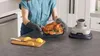 A person is holding a roasted chicken in a glass dish, with lemons and a knife on a cutting board in the foreground, and a kitchen appliance in the background. Anteprima dell’immagine prodotto