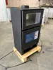 A black modern wood-burning stove is displayed on a wooden pallet in a warehouse or storage area. Produktbild-Vorschau