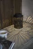 A black metal lantern with a leaf pattern is placed on a surface, casting leaf-shaped shadows. A white coffee cup and a magazine are also visible. Produktbild-Vorschau