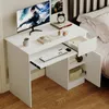 A white computer desk is shown next to a bed, with a monitor, keyboard, mouse, books, and headphones on it. The desk has a drawer and a cabinet with a shelf. Podgląd zdjęcia produktu