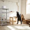 A person is working at a workbench in a workshop, with shelves of supplies to the left. Produktbild-Vorschau